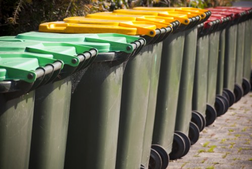 A row of large, modern wheelie bins used for rubbish collection, positioned on a paved outdoor surface. The bins are made of durable plastic with smooth, slightly glossy finishes, and are arranged in a line. The frontmost bins are green with black wheels, followed by yellow-lidded bins, and then red-lidded bins further along the row. The lids are securely closed, and the bins are placed close together, extending into the background along a residential street. The surrounding environment features a pavement with some small patches of grass visible at the base of the bins, and natural daylight illuminates the scene. The image captures waste management and rubbish removal services in a typical local setting, related to companies such as St Johns Wood House Clearance operating in nearby areas like St Johns Wood.