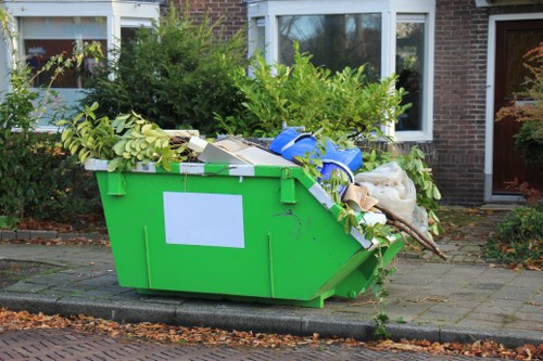 A large, bright green waste skip filled with assorted garden and household rubbish, situated on a paved driveway outside a residential property in a suburban area. The skip contains thick branches with green leaves, cardboard boxes, plastic containers, bags, and wooden items, with some materials spilling over the top edge. The background features a brick house with white-framed windows and a brown door, surrounded by shrubbery and small trees, indicating a quiet neighbourhood likely within or near St Johns Wood. The scene is lit with natural daylight, creating clear visibility of the various textures and colors of the discarded items, consistent with a rubbish removal service such as offered by St Johns Wood House Clearance.