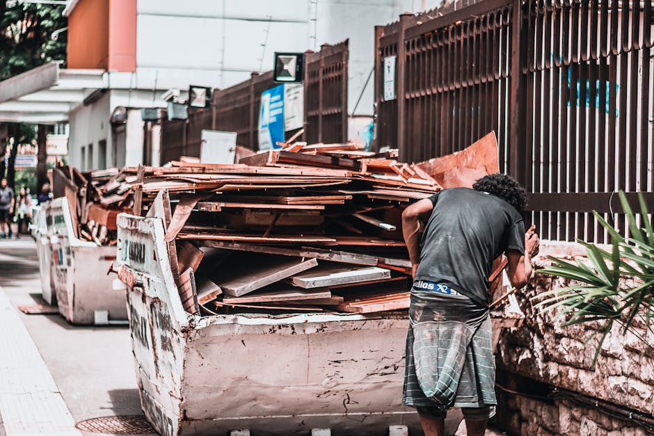 A man with curly dark hair, dressed in a black t-shirt and grey plaid shorts, is seen from the back standing next to a large, overflowing skip filled with construction debris such as wooden planks, ti
